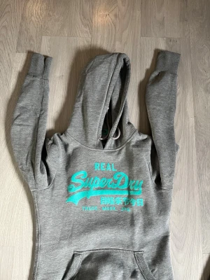 Grå Superdry hoodie med turkost tryck - Snygg grå hoodie från Superdry med turkost tryck på bröstet och japanska tecken. Klassisk känguruficka framtill och huva med dragsko. Mjuk bomullsblandning och ribbade muddar vid ärmar och nederkant. Perfekt för en avslappnad streetwear-look.