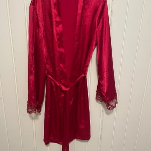 Röd satin morgonrock med spetsdetaljer - Röd morgonrock i glansigt satinmaterial med knytskärp i midjan och spetsdetaljer vid ärmsluten. Perfekt för en lyxig känsla hemma. Långärmad och lätt i materialet.