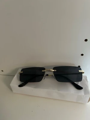 Svarta rektangulära solglasögon - Snygga solglasögon med rektangulära, svarta glas och tunna guldfärgade bågar. Minimalistisk design utan synlig ram runt glasen och svarta skalmar. Perfekta för dig som vill ha en clean och modern look.