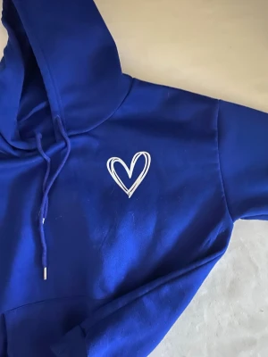 Blå hoodie med vitt hjärta - Snygg blå hoodie med huva och dragsko, samt en vit hjärt-detalj tryckt på bröstet. Hoodien har en klassisk känguruficka framtill och är gjord i mjukt material, perfekt för chill dagar. Passar dig som gillar enkel men stilren streetwear.pris kan diskuteras