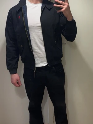 Svart jacka från Polo Ralph Lauren - Snygg svart jacka från Polo Ralph Lauren med klassisk krage, dragkedja och knappar. Jackan har två fickor framtill och den ikoniska röda loggan broderad på bröstet. Materialet är bomull och jackan har en stilren, tidlös look.