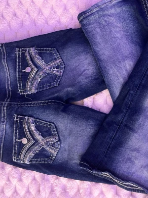 Mörkblå jeans med broderade fickor - Säljer ett par mörkblå jeans med snygga slitningar och broderade detaljer på bakfickorna. Jeansen har kontrastsömmar och dekorativa knappar på ficklocken. Perfekta för dig som gillar unika detaljer och en trendig look.