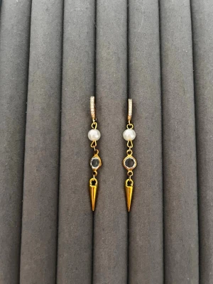 Guldtonade örhängen med pärlor och spets - Säljer ett par unika örhängen i guldton med vita pärlor, genomskinliga stenar och spetsiga hängen. Örhängena har en cool kombination av material och former som ger en edgy men elegant vibe. Perfekta för dig som vill sticka ut lite extra.