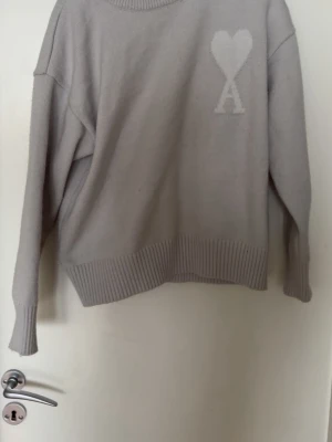stickad tröja Ami Paris - Ljusgrå stickad tröja från Ami Paris med ikoniskt hjärta och A-logga på bröstet. Tröjan har rund halsringning, ribbade muddar och nederkant. Tillverkad i mjukt material som ger en avslappnad och trendig look.