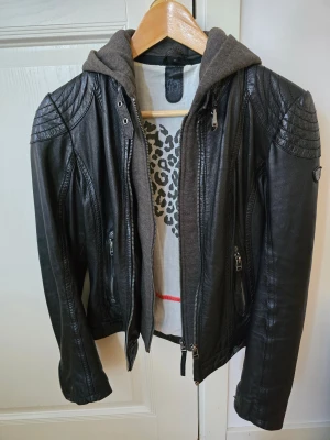 Svart skinnjacka med grå luva - XS - Snygg svart skinnjacka med grå luva och coola detaljer på axlarna. Jackan har dragkedjor framtill och på fickorna samt en figurnära passform. Perfekt för dig som vill ha en edgy look med streetkänsla. Knappt använd, nästan nyskick bortsett från mindre reva, se bild. 100% läder. Luvan är avtagbar. Nypris 1800:-