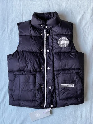 Svart dunväst från Canada Goose - Snygg svart dunväst från Canada Goose med hög krage, två stora framfickor och tydlig logotyp på bröstet. Västen har både dragkedja och tryckknappar framtill samt en klassisk puffy design. Perfekt för lager-på-lager och streetstyle.