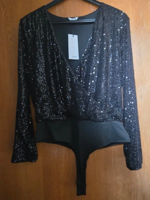Svart paljettbody från mbyM - Snygg svart body från mbyM med långa ärmar och djup omlott-ringning. Överdelen är täckt av glittrande paljetter som ger en festlig look, medan underdelen är i slätt tyg. Perfekt för dig som vill sticka ut lite extra. Materialet är en mix av polyester och elastan.