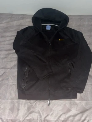 Svart Nike hoodie med dragkedja - Säljer en svart hoodie från Nike med dragkedja framtill och två fickor med dragkedja. Klassisk huva och gul Nike-logga på bröstet. Perfekt för en sportig och avslappnad stil. Tillverkad i mjukt material för extra komfort.