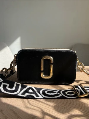 Svart Marc Jacobs Snapshot handväska - Säljer en svart Marc Jacobs Snapshot väska i fint skick. Perfekt accessoar för dig som gillar trendiga detaljer.