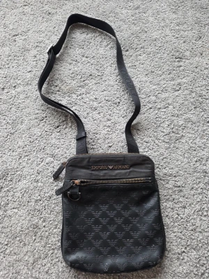 Svart axelväska Emporio Armani - Cool svart axelväska från Emporio Armani med justerbar rem och snyggt mönster av logotypen på framsidan. Väskan har två dragkedjefack och är tillverkad i slitstarkt syntetmaterial. Perfekt för att bära dina essentials och ge din outfit en lyxig touch.