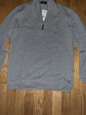 Grå halvzip från Polo Ralph Lauren - Snygg grå tröja med halv dragkedja från Polo Ralph Lauren. Klassisk passform med broderad logga på bröstet. Perfekt för lager-på-lager och enkel att matcha med jeans eller chinos.