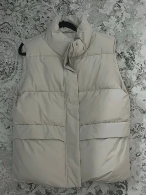 Beige dunväst från Dobber - Snygg beige dunväst från Dobber med hög krage och breda kanaler. Västen har dold dragkedja och tryckknappar framtill samt två stora fickor. Skick 10/10 aldrig använt. Storlek xs men oversize, passar S-M. Nypris: 500 kr