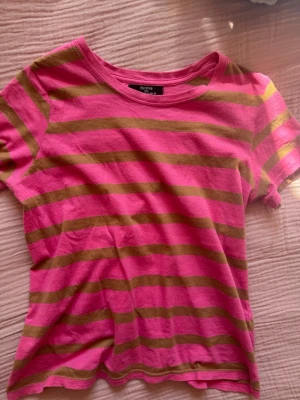 Randig rosa t-shirt från Bershka - Säljer en kortärmad t-shirt från Bershka i rosa med olivgröna ränder. T-shirten har en klassisk rund halsringning och är gjord i mjuk bomull, perfekt för en chill och färgglad look.