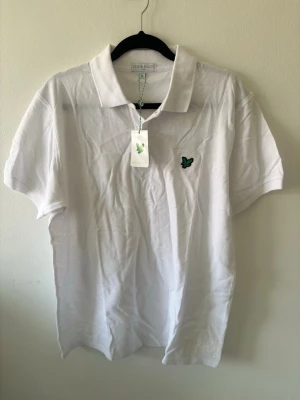 Vit pikétröja från Lyle & Scott. Passar även L  - Klassisk vit pikétröja från Lyle & Scott med korta ärmar och krage. Tröjan har en broderad logga i grönt och svart på bröstet samt knappar vid halsen. Perfekt till en avslappnad men stilren look.