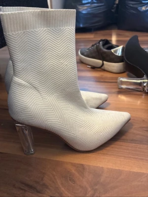 Vita stickade stövletter med plexiklack - Säljer ett par vita stövletter med stickad struktur i sicksackmönster och spetsig tå. Skorna har en genomskinlig plexiklack som ger en modern touch. Materialet är stretchigt tyg som sitter tight runt ankeln. Perfekta för dig som vill ha något trendigt och unikt. De är smutsiga pga skor som legat över dem, kan torkas av med trasa