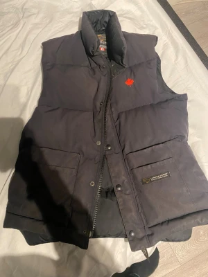 Svart dunväst från Canada Goose - Snygg svart dunväst från Canada Goose med hög krage och två stora framfickor. Västen har en röd lönnlöv-detalj på bröstet och klassisk Canada Goose-logga på fickan. Perfekt för lager-på-lager och håller dig varm under kyliga dagar. Storlek S