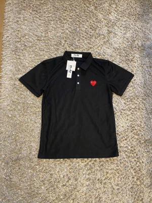 Svart piké från Comme des Garçons Play - Svart pikétröja från Comme des Garçons Play med klassisk krage, knappar framtill och det ikoniska röda hjärtat broderat på bröstet. Kortärmad och i mjuk bomull, perfekt för en clean och stilren look.