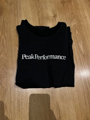 Svart Peak Performance t-shirt - Snygg svart t-shirt från Peak Performance med klassisk rund hals och vit logga tryckt framtill. Tillverkad i mjukt bomullsmaterial som känns skönt mot huden. Perfekt till jeans eller shorts för en clean och enkel stil.