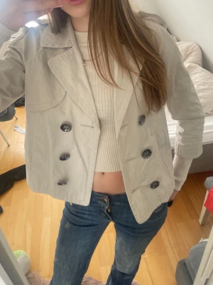 Beige kort trenchcoat med stora knappar - skit Snygg beige trekvartsärmad trenchcoat med dubbelknäppning och stora svarta knappar. Jackan har bred krage och är lite kortare i modellen, passar perfekt att ha till ett par jeans och en långärmad tröja som på bilderna. jackan är från Nafnaf och är i storlek s men skulle säga att den sitter som en xs-xxs. älskar den här jackan  men den har blivit för liten för mig. den är i super bra skick. hör av dig om du har några frågor eller funderingar 💕