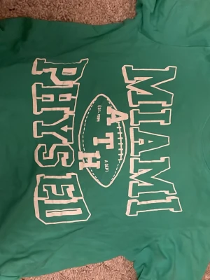 Grön Miami Phys Ed T-shirt - Snygg grön t-shirt med stort vitt tryck där det står 'MIAMI PHYS ED' och en amerikansk fotboll i mitten. Perfekt för dig som gillar sportig stil och college-vibe. Klassisk passform och rund hals.