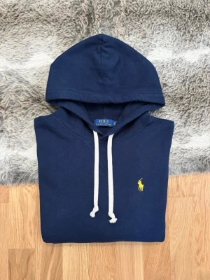 Marinblå Hoodie Från Polo Ralph Lauren - Storlek XS - Tjena!👋 Säljer en riktigt snygg marinblå Polo Ralph Lauren hoodie i storlek XS (Herr). Den har den klassiska pony-loggan broderad i gult på bröstet – en grym färgkombination! 🐎 Tröjan är i mycket fint skick, välvårdad och har kvar sin mjuka fleece-känsla på insidan ✨ Sitter perfekt för dig som drar XS eller vill ha en mer slimmad fit 👕 Ingen QR-kod på denna, men alla tvättlappar sitter kvar och garanterar äktheten ✅ Ett stilrent och lyxigt basplagg. Fraktar snabbt! 🚛📦💬💯 