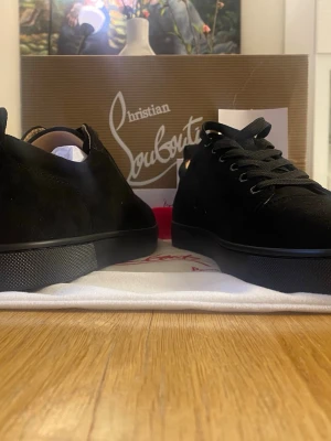 Svarta sneakers från Christian Louboutin - Snygga svarta sneakers från Christian Louboutin i mocka med klassisk röd sula. Skorna har en låg profil, svarta skosnören och diskret design som ger en exklusiv känsla. Perfekta för dig som gillar stilrena och lyxiga detaljer.