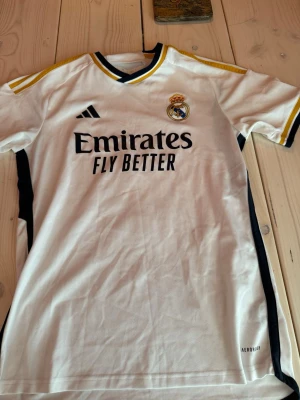 Real Madrid matchtröja Bellingham 5 - Säljer en vit Real Madrid matchtröja från Adidas med svarta och gula detaljer. Tröjan har klubbmärke, sponsortryck och Bellingham 5 på ryggen. Tillverkad i lätt och ventilerande material med Aeroready-teknologi. Perfekt för fotbollsträning eller att bära till vardags.