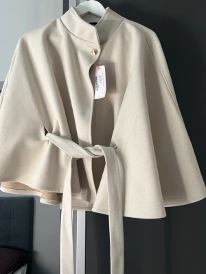 Beige cape-kappa med knytband - Stilren beige cape-kappa med hög krage, knytband i midjan och knappstängning upptill. Kappan har en elegant, vid siluett och är tillverkad i ett mjukt tyg som ger ett exklusivt intryck. Perfekt för dig som vill ha en trendig och tidlös look.