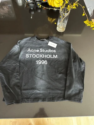 Svart långärmad topp Acne Studios - Cool svart långärmad topp från Acne Studios med vit text framtill där det står 'Acne Studios STOCKHOLM 1996'. Klassisk rund halsringning och relaxed fit. Perfekt för dig som gillar streetwear och vill ha något stilrent men ändå statement.