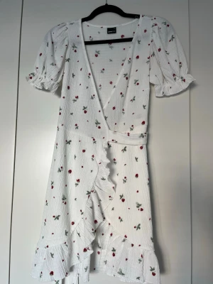 Vit omlottklänning med blommor - Söt vit omlottklänning med röda blommor och gröna blad, volangkant och puffiga kortärmar med resår. Klänningen har knytband i midjan och är tillverkad i ett lätt, strukturerat tyg som passar perfekt till sommaren.