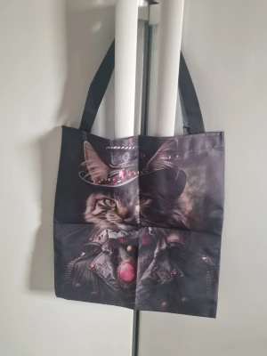 Tygväska kattmotiv kvinna - Tygväska för kvinna. Väskan är tillverkad i polyestertyg och har tryck med cool katt framsidan och baksidan. Utrustad med två axelremmar i kontrastfärg. Passar för vardagsbruk, till exempel som shoppingväska eller extra vardagsväska. 33,5 cm x 38 cm.