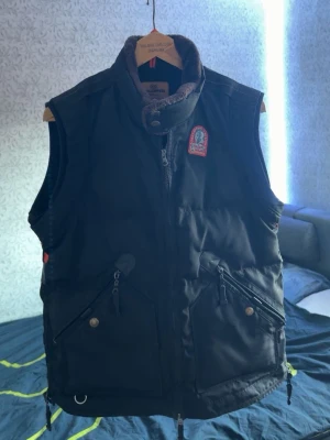 Svart dunväst från Parajumpers - Svart dunväst från Parajumpers med hög krage och dragkedja framtill. Västen har två stora fickor med dragkedja och tryckknapp samt en ikonisk Parajumpers-logga på bröstet. Materialet är quiltat och fyllt med dun för extra värme, den är i väldigt bra skick o har inga hål eller slitage om du har någon mer fråga så är du välkommen i pm! Mvh 