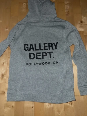 Grå hoodie från Gallery Dept. - Snygg grå hoodie från Gallery Dept. med stor svart text på ryggen: 'GALLERY DEPT. HOLLYWOOD, CA.'. Klassisk passform med huva och känguruficka framtill. Tillverkad i mjukt sweatshirtmaterial, perfekt för en avslappnad streetwear-look.