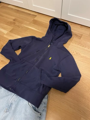 Mörkblå hoodie från Polo Ralph Lauren - Snygg mörkblå hoodie från Polo Ralph Lauren med dragkedja framtill och klassisk gul logga på bröstet.  Hoodien är storlek s men passar som en xs