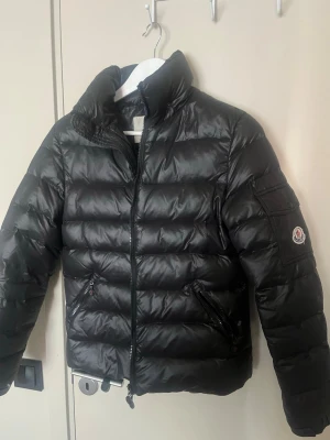 Svart dunjacka från Moncler - Snygg svart dunjacka från Moncler med hög krage och två dragkedjefickor framtill. Jackan har quiltad design och Moncler-logga på ärmen. Tillverkad i glansigt material som ger en exklusiv look. Perfekt för en sen lite kallare sommarkväll eller hösten 😍