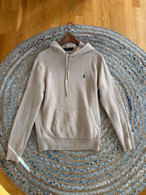 Beige hoodie från Polo Ralph Lauren - Säljer en beige hoodie från Polo Ralph Lauren med klassisk huva och dragsko. Tröjan har ett diskret broderat logotyp på bröstet och en stor magficka. Stickad struktur och ribbade muddar vid ärmslut och nederkant. Perfekt för en chill look.