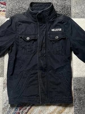 Mörkblå Fieldjacket från Hollister - Cool mörkblå Fieldjacket från Hollister med hög krage och två bröstfickor med knappar. Jackan har en rak passform och är tillverkad i ett slitstarkt bomullsmaterial. Hollister-loggan sitter på vänster bröst. Perfekt för att ge din outfit en avslappnad vibe.
