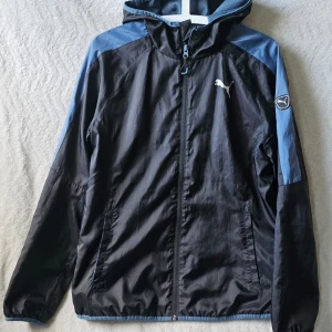 Svart och blå vindjacka från Puma - Snygg vindjacka från Puma i svart med blå detaljer på axlar och ärmar. Jackan har huva, dragkedja framtill och två sidofickor. Puma-logga på bröstet och ärmen. Materialet är lätt och perfekt för att skydda mot vind.