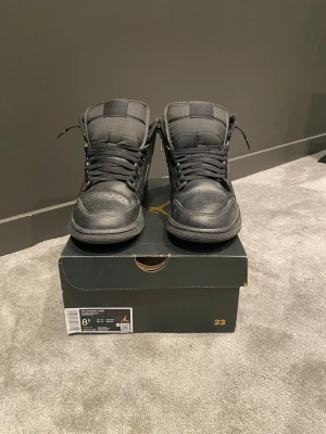 Svarta Air Jordan 1 Mid sneakers - Säljer ett par helsvarta Air Jordan 1 Mid sneakers i läder med klassisk siluett och svarta skosnören. Skorna har vadderad plös och krage samt diskret Jumpman-logga på plösen. Perfekta för dig som gillar en klassisk och simpel look. Snörena är längre men jag har stoppat in dom i skon för ett bättre utseende. Det är ett litet hål på kanten av skolådan som visas på bild men inget märkvärdigt.