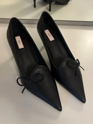 Svarta pumps med rosett - Stilrena svarta pumps från Nelly med spetsig tå och liten rosett framtill. Helt oanvända.