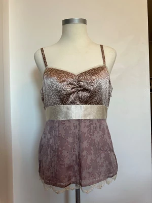 Lila och beige spetsbody med mönster - Söt body i lila och beige med tunna axelband och blommigt mönster upptill. Midjan markeras av ett brett, glansigt band i satin och nederdelen har ett skirt tyg med blommig spetskant. Materialet är tunt och glansigt, troligen polyester eller satinblandning.