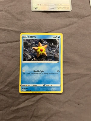 Pokémonkort Staryu - Säljer ett Pokémonkort med Staryu-motiv. Kortet har blå och gul ram, med en bild på Staryu mot en stenig bakgrund. Text och detaljer på engelska. Perfekt för samlare eller Pokémonfans.
