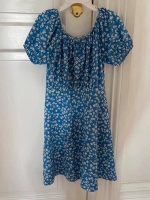 Blå blommig kortklänning - Söt blå kortklänning med vitt blommönster och puffiga korta ärmar. Klänningen har en rundad halsringning och lätt markerad midja. Perfekt för sommarens alla tillfällen och ger en fräsch och ungdomlig look.