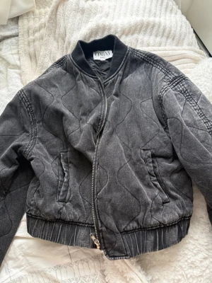 Svart quiltad bomberjacka CIMINY - Snygg svart quiltad bomberjacka från CIMINY Paris. Jackan har ribbade muddar, dragkedja framtill och två fickor. Materialet är mjukt och har ett vågigt quiltat mönster som ger en cool vintagekänsla. Perfekt för dig som vill ha en unik och trendig look.