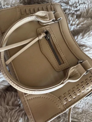 Beige handväska från Gina Tricot - Snygg beige handväska från Gina Tricot i konstläder med dekorativa sömmar och flätade detaljer. Väskan har både handtag och axelrem, samt en praktisk ytterficka med dragkedja och flera fack inuti. Perfekt för dig som gillar stilrena accessoarer.