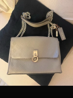 GUESS handväska  - Snygg grå handväska från Guess med präglat logomönster över hela väskan. Väskan har silverfärgade metalldetaljer, kedjerem och ett dekorativt Guess-hänge. Som ni ser har färgen släppt på två ställen, inget som syns men går att åtgärda för den som har tid. Man skulle kunna ta bort dem hängerna o sätta på nästa. 