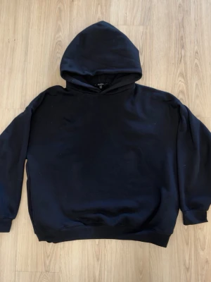 Svart basic hoodie unisex oversized - En enkel svart hoodie med huva och långa ärmar. Klassisk modell utan tryck eller detaljer, perfekt för en clean och avslappnad stil. Tillverkad i mjukt bomullsmaterial som känns skönt mot huden. Passar både killar och tjejer.