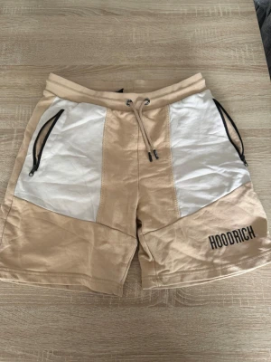 Beige och vita shorts från Hoodrich - Snygga beige shorts från Hoodrich med vita paneler framtill och svarta dragkedjor på fickorna. Shortsen har snörning i midjan och en bakficka med Hoodrich-logga. Perfekta för sommaren och avslappnad stil.