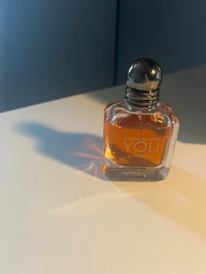 Stronger With You Intensely parfym - Stronger With You Intensely från Emporio Armani är en elegant parfym i en fyrkantig glasflaska med rundad silverfärgad kork. Doften har en varm bärnstensfärgad vätska och flaskan har stilren design med tydlig logotyp framtill.