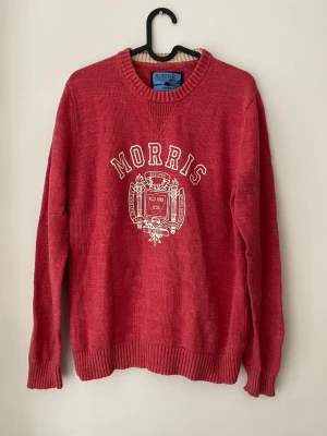 Röd stickad tröja från Morris - Röd stickad tröja från Morris med vitt broderat emblem och text på bröstet. Tröjan har rund halsringning, ribbade muddar vid ärmslut och nederkant samt lång ärm. Perfekt för dig som gillar klassisk college-stil.